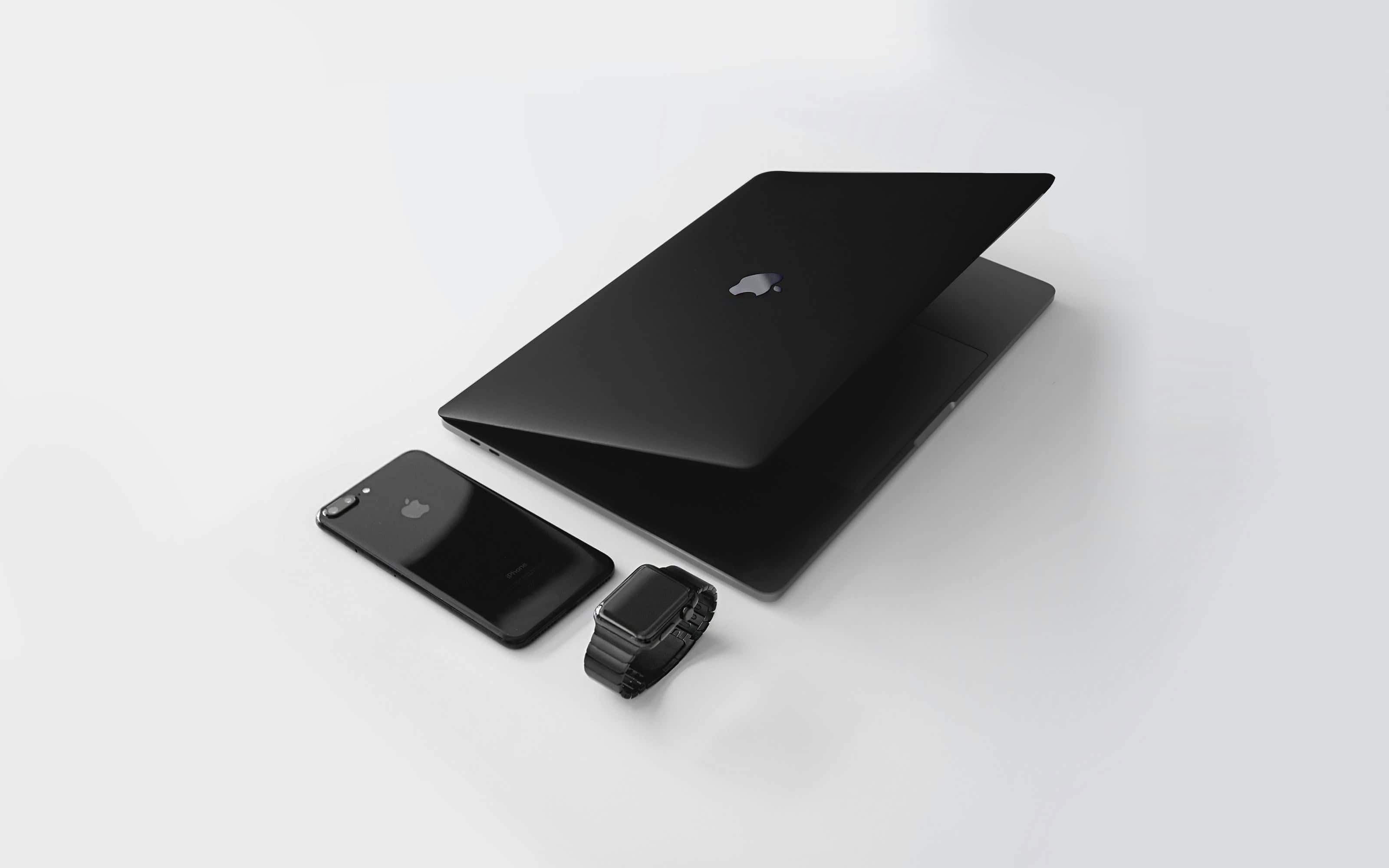 Kuvassa Apple laitteet, musta Macbook pro, musta iPhone suojakotelolla, sekä musta Apple Watch